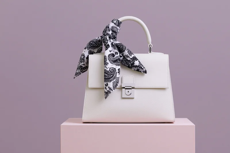 White structured leather handbag with silver clasp on pink podium against lavender background