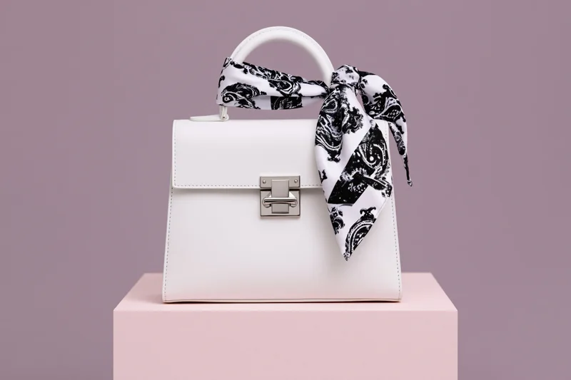White structured leather handbag with silver clasp and black-and-white paisley scarf on pink podium