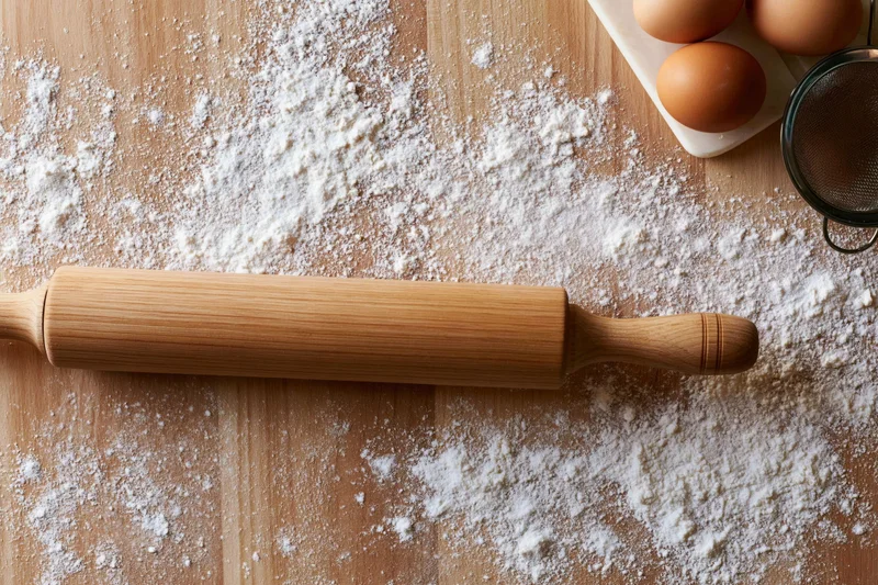 Wooden rolling pin on flour-dusted kitchen countertop with eggs and sifter nearby