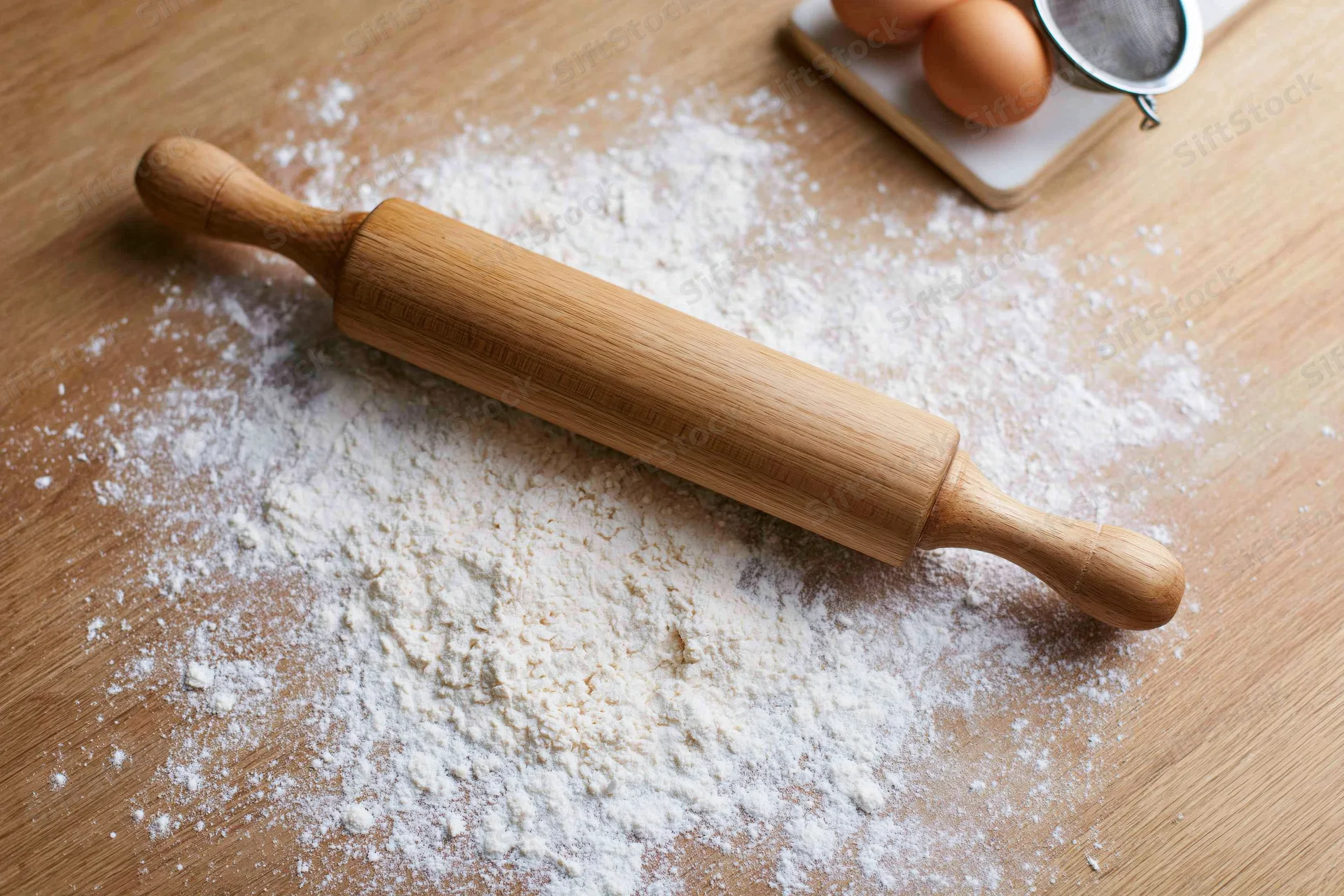 Wooden rolling pin resting on flour-dusted kitchen countertop with eggs and sifter nearby