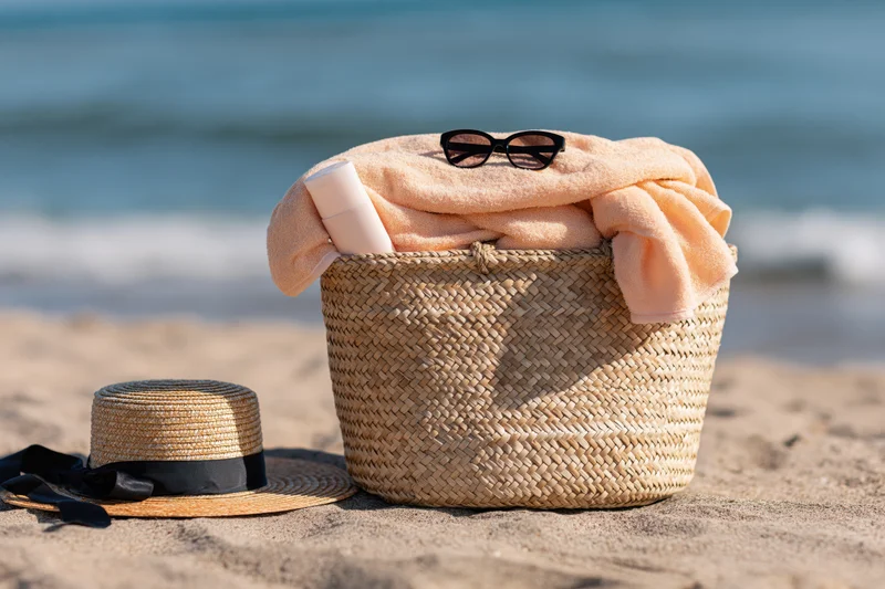 Woven straw basket filled with peach towel and sunglasses on sandy beach near ocean