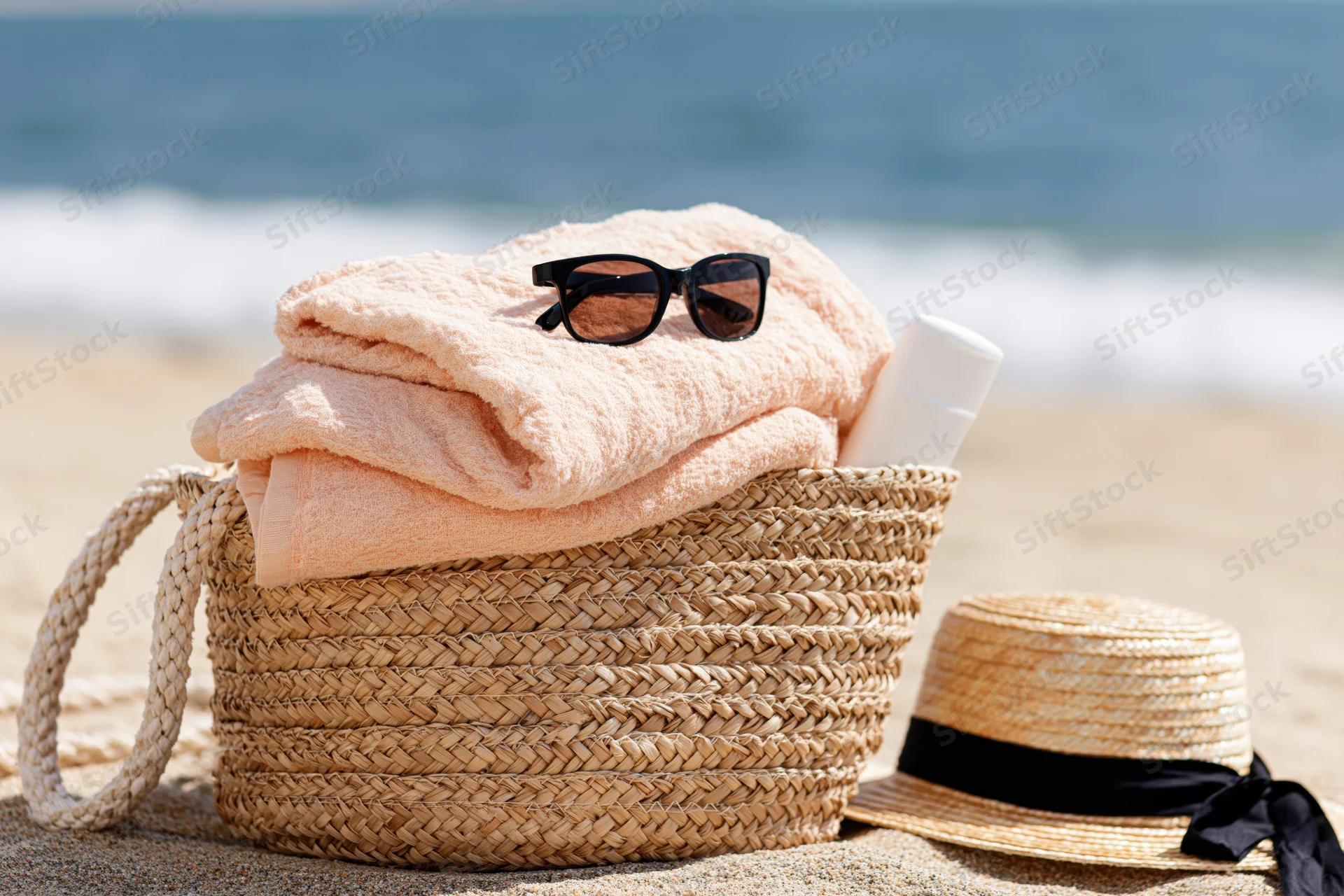 Woven straw beach bag with peach towel, sunglasses and hat on sandy shore near ocean