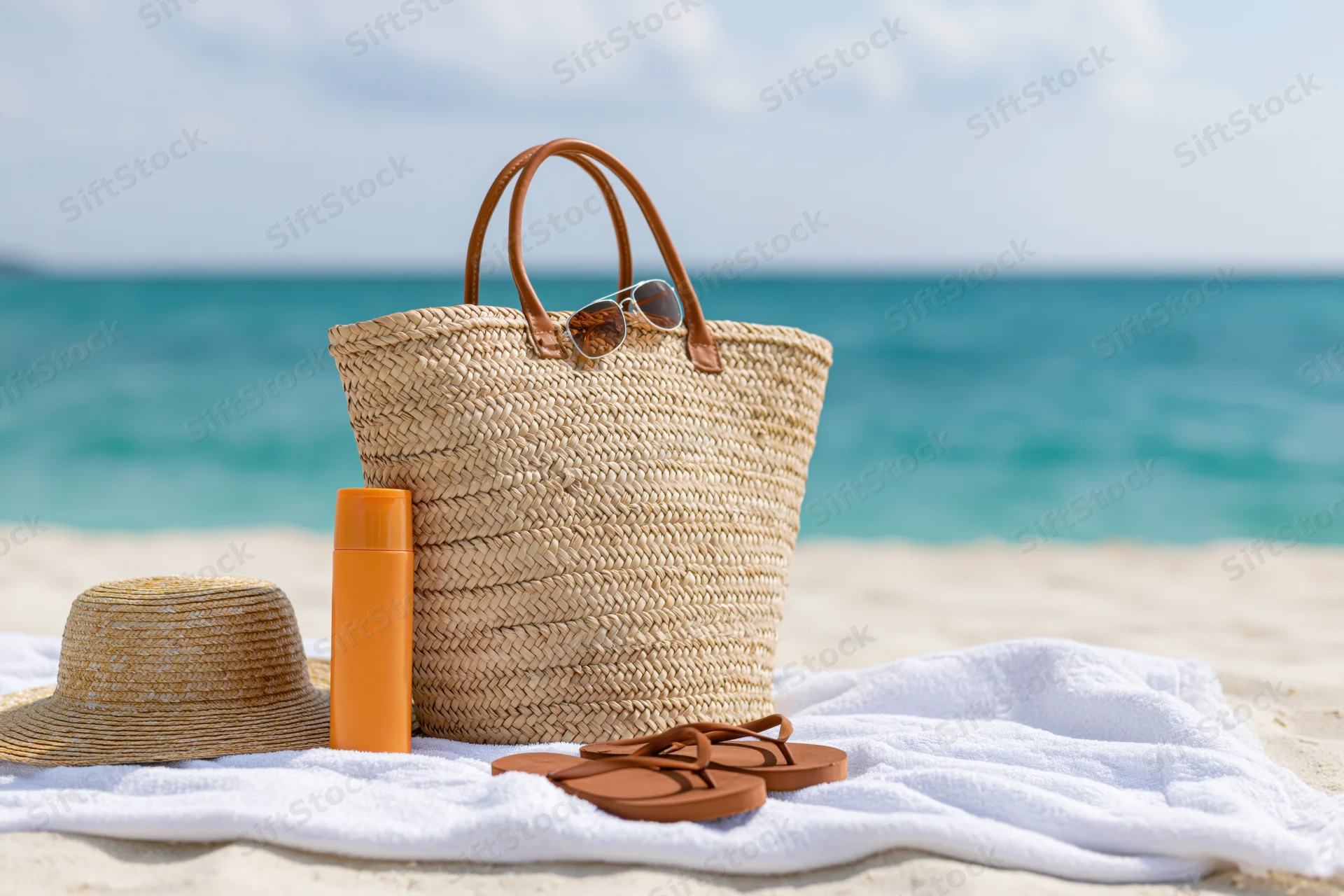 Woven straw beach bag with sunglasses and accessories on towel by ocean