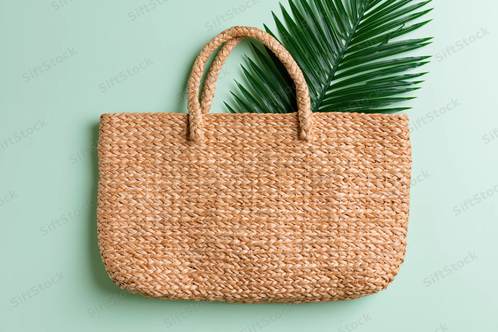 Woven straw tote bag with palm leaf on mint green background