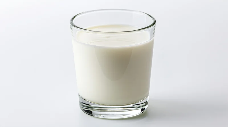 A clear glass tumbler filled with milk on a white surface