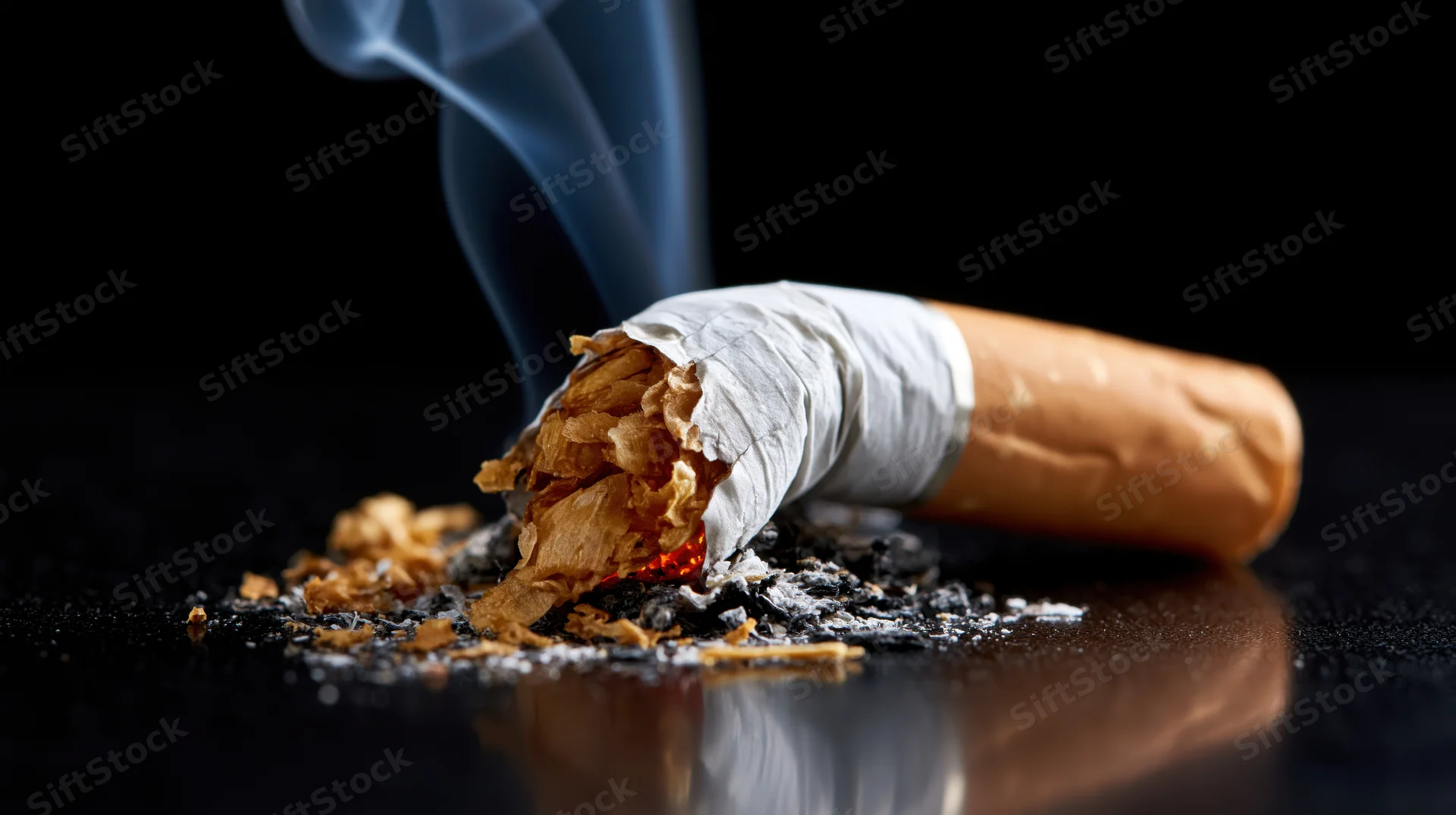 Crushed cigarette butt with smoke and ash on a dark reflective surface
