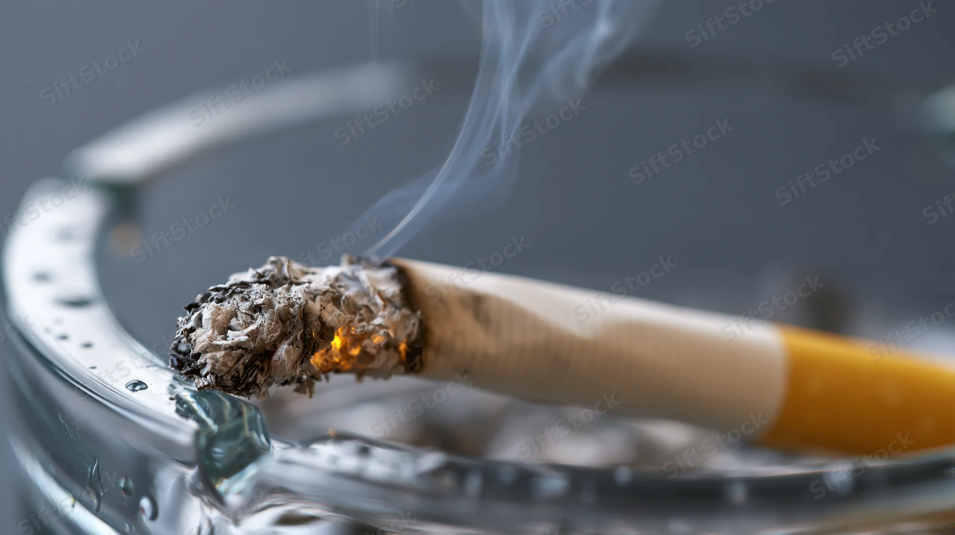 Lit cigarette with glowing ember and smoke resting in a glass ashtray