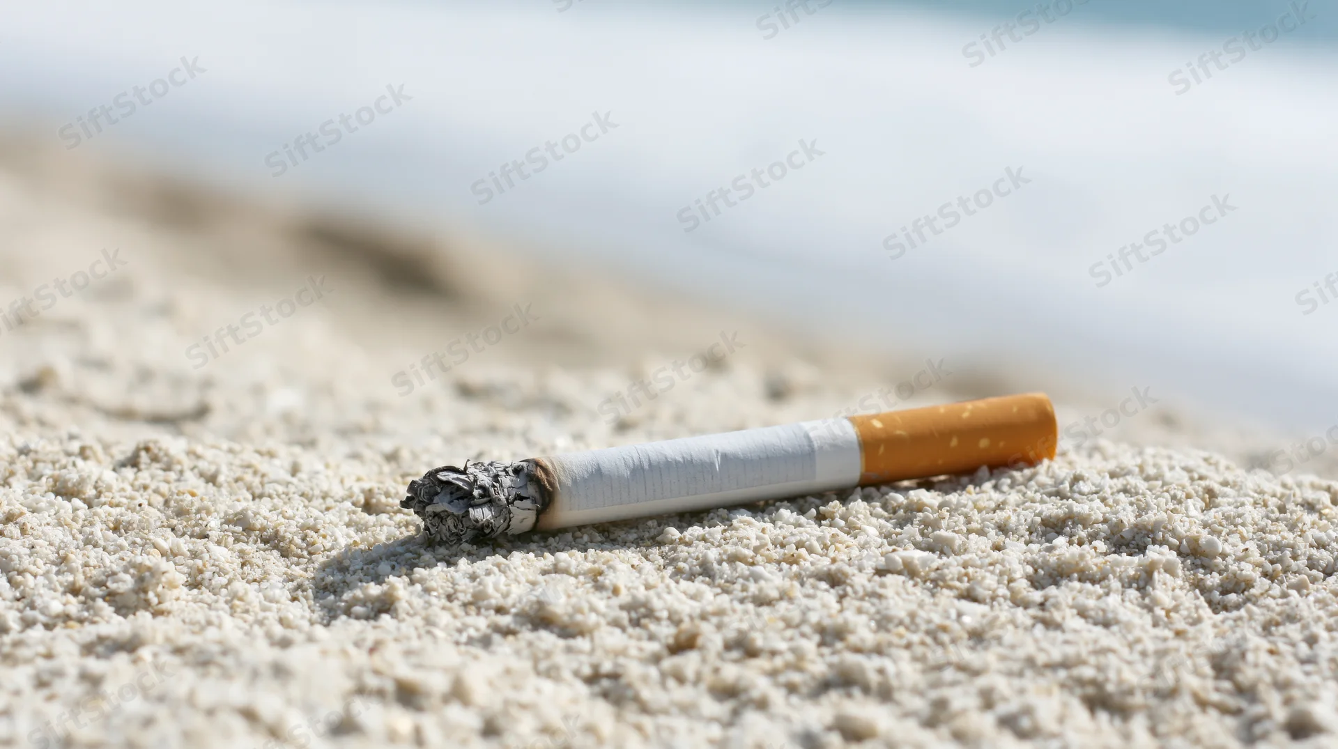Lit cigarette lying on coarse white sand at beach with ocean in background