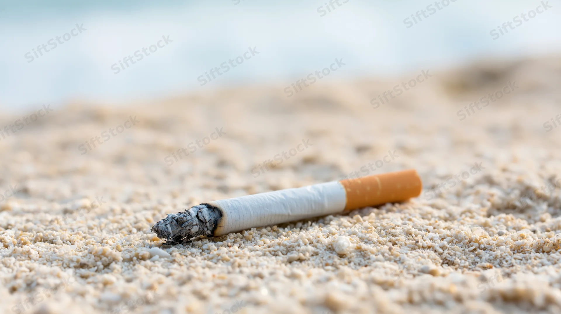 Lit cigarette lying on coarse white sand at beach with ash and blurred ocean background