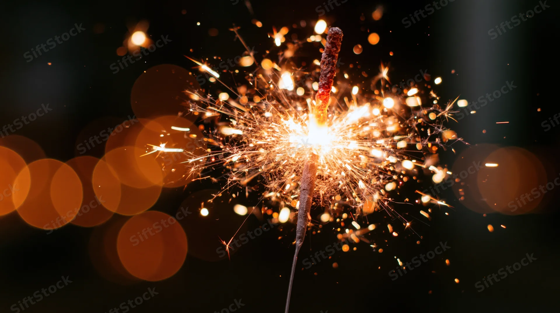 Lit sparkler burning with sparks flying outward against dark background with bokeh lights