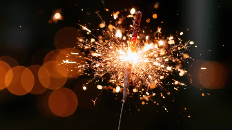 Lit sparkler burning with sparks flying outward against dark background with bokeh lights