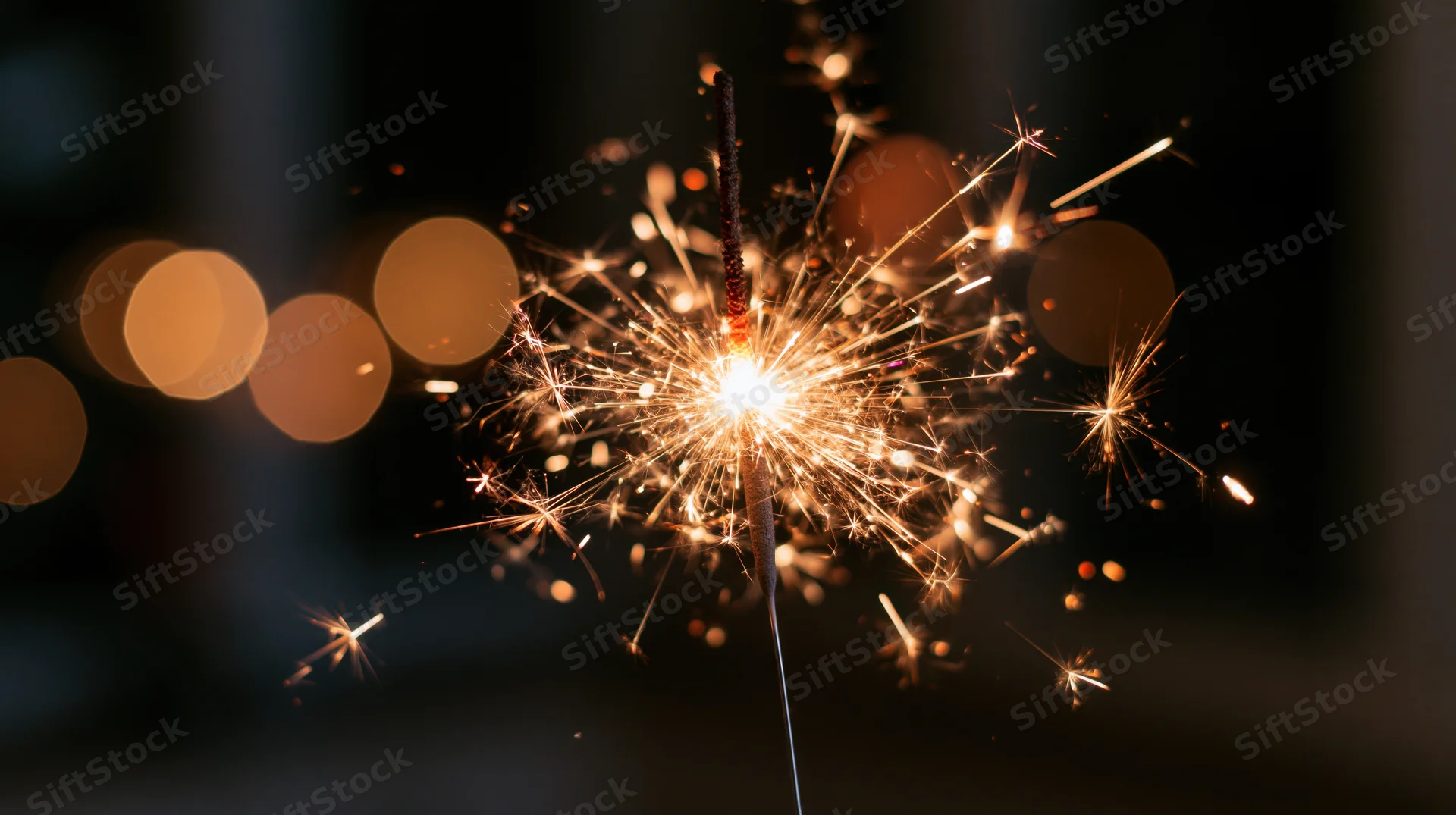 Lit sparkler burning with sparks flying outward against dark background with bokeh lights