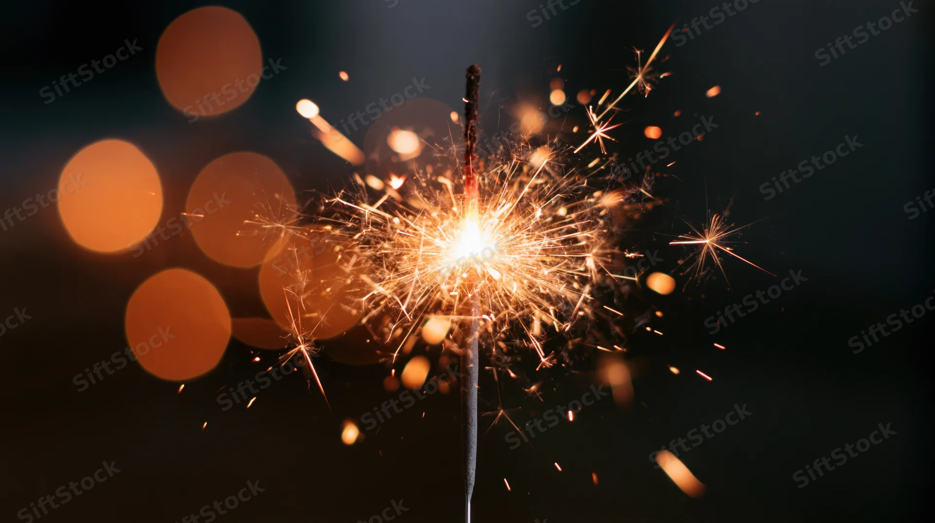 Lit sparkler burning with sparks flying outward against dark background with bokeh lights
