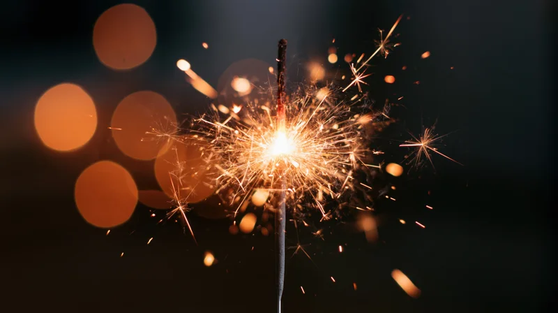 Lit sparkler burning with sparks flying outward against dark background with bokeh lights