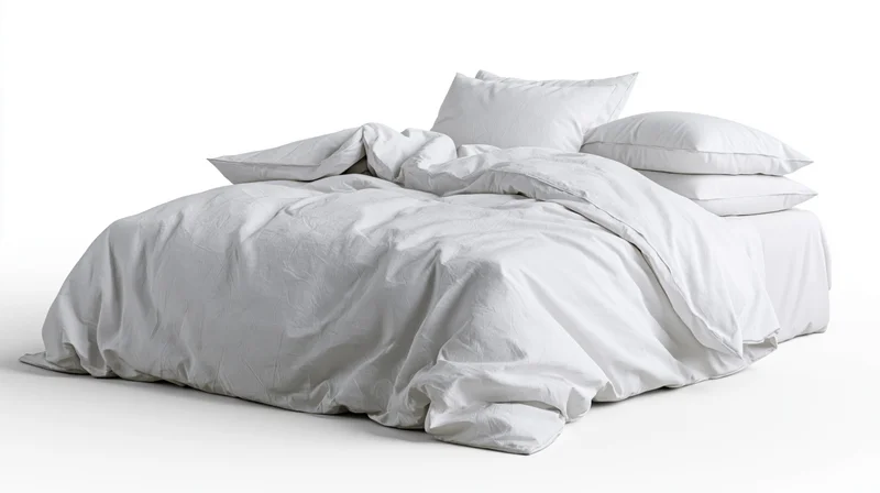 Unmade bed with rumpled white cotton duvet and stacked pillows on plain white background