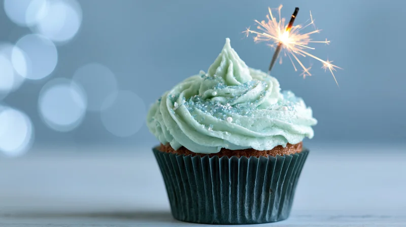 Single birthday cupcake with swirled mint-blue frosting and lit sparkler on a light surface with bokeh background