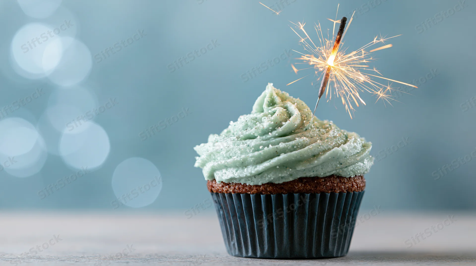 Single birthday cupcake with lit sparkler on mint frosting against blurred blue background