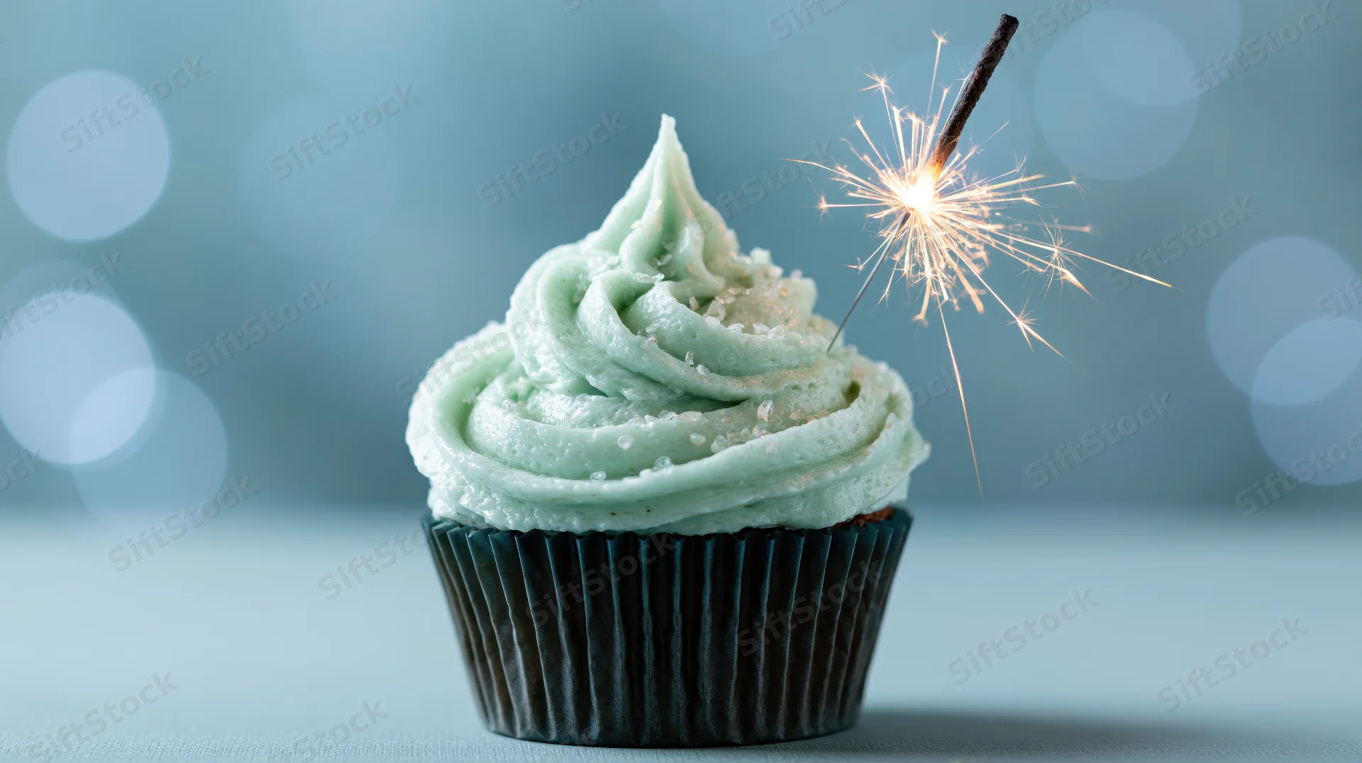 Single birthday cupcake with swirled mint frosting and lit sparkler on blue background