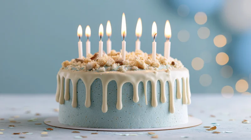 Blue birthday cake with white drip icing and nine lit candles on a table with confetti and bokeh lights in the background