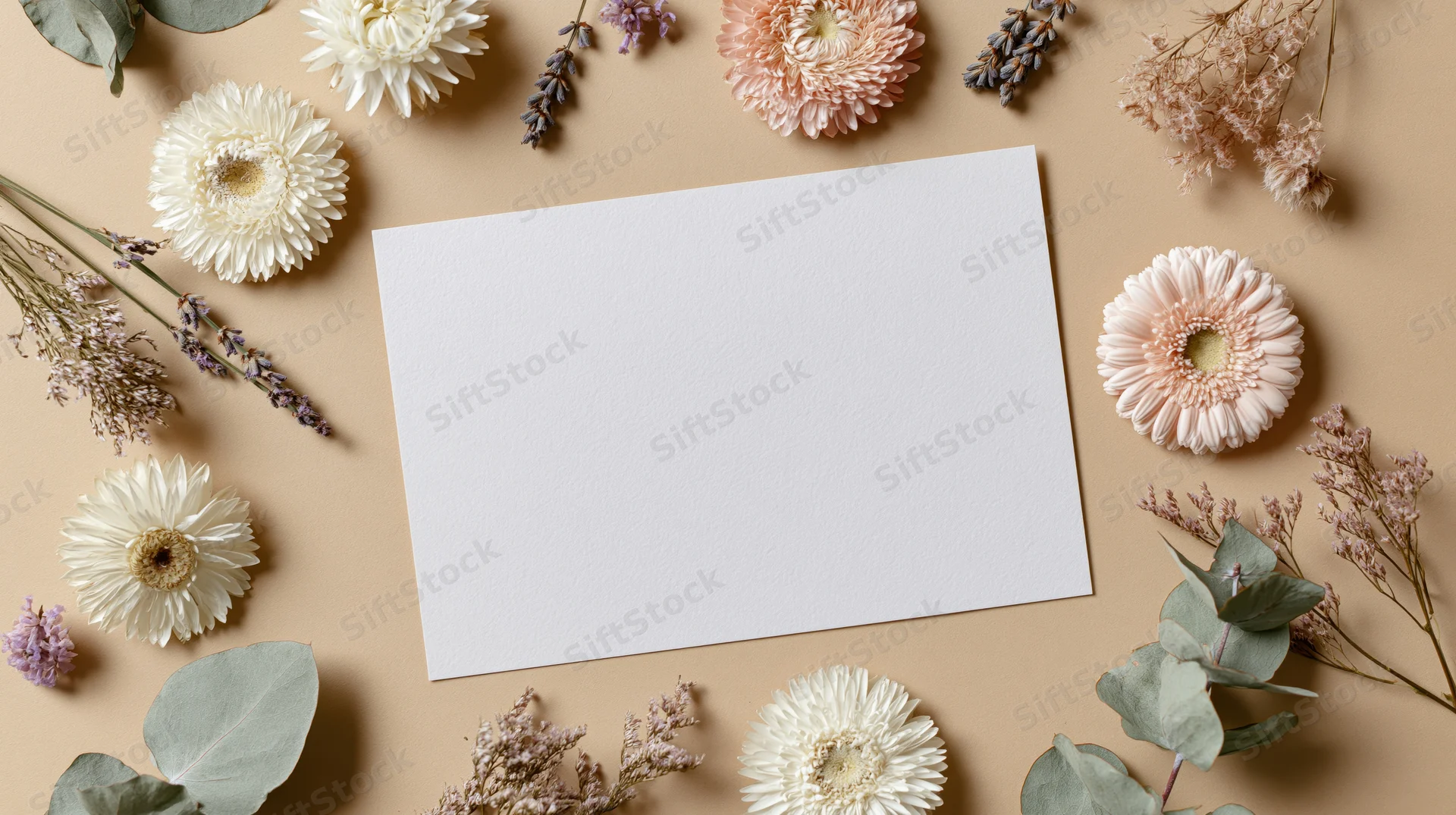 Blank white card surrounded by dried flowers and greenery on beige surface