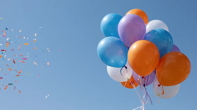 Cluster of orange blue purple and white helium balloons floating in clear blue sky with confetti drifting nearby