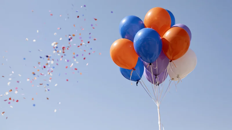Cluster of orange blue purple and white helium balloons floating in sky with confetti drifting nearby