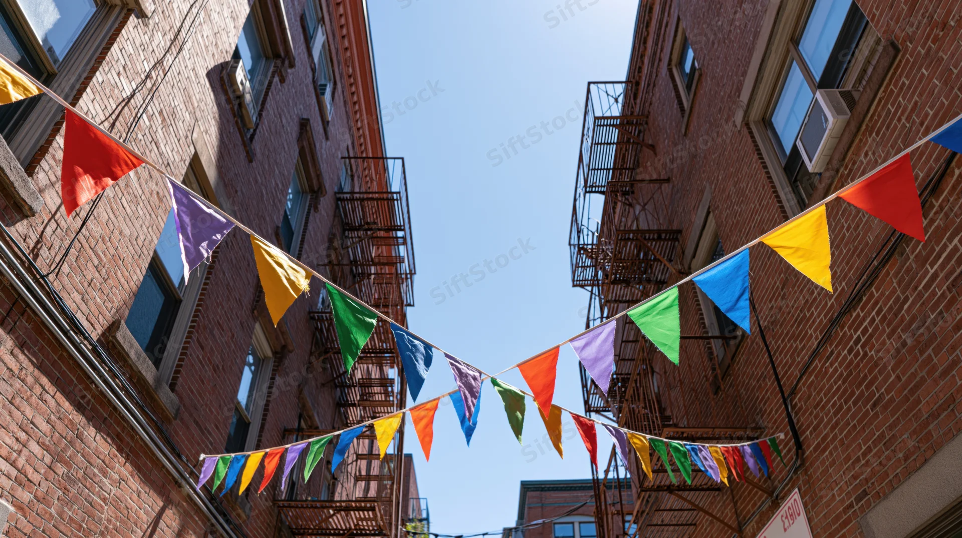 Colorful triangular bunting flags strung across a narrow urban alley between brick buildings with fire escapes under a clear blue sky