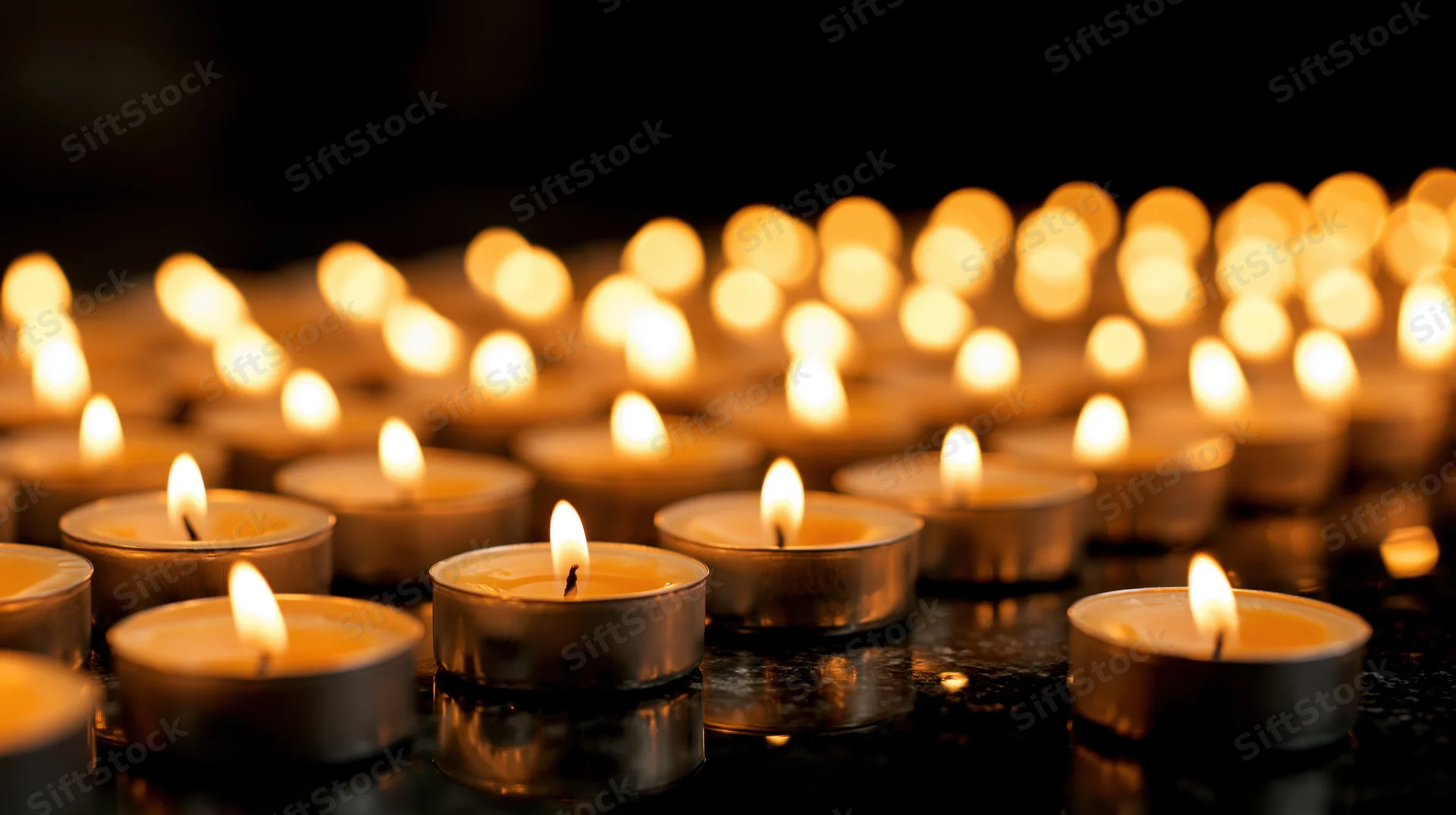 Dozens of small silver tealight candles burning in rows on a dark reflective surface