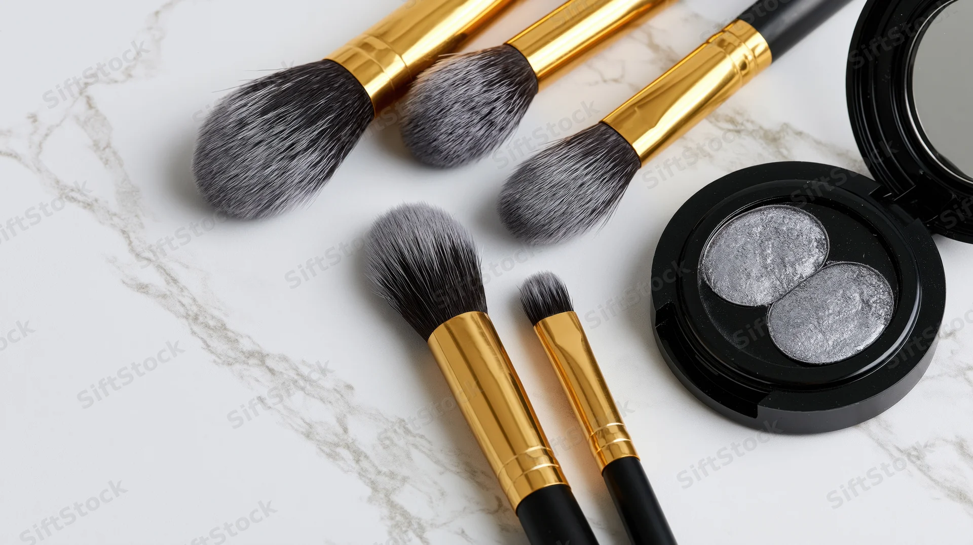 Makeup brushes with gold ferrules and black handles arranged with a silver eyeshadow compact on a white marble surface