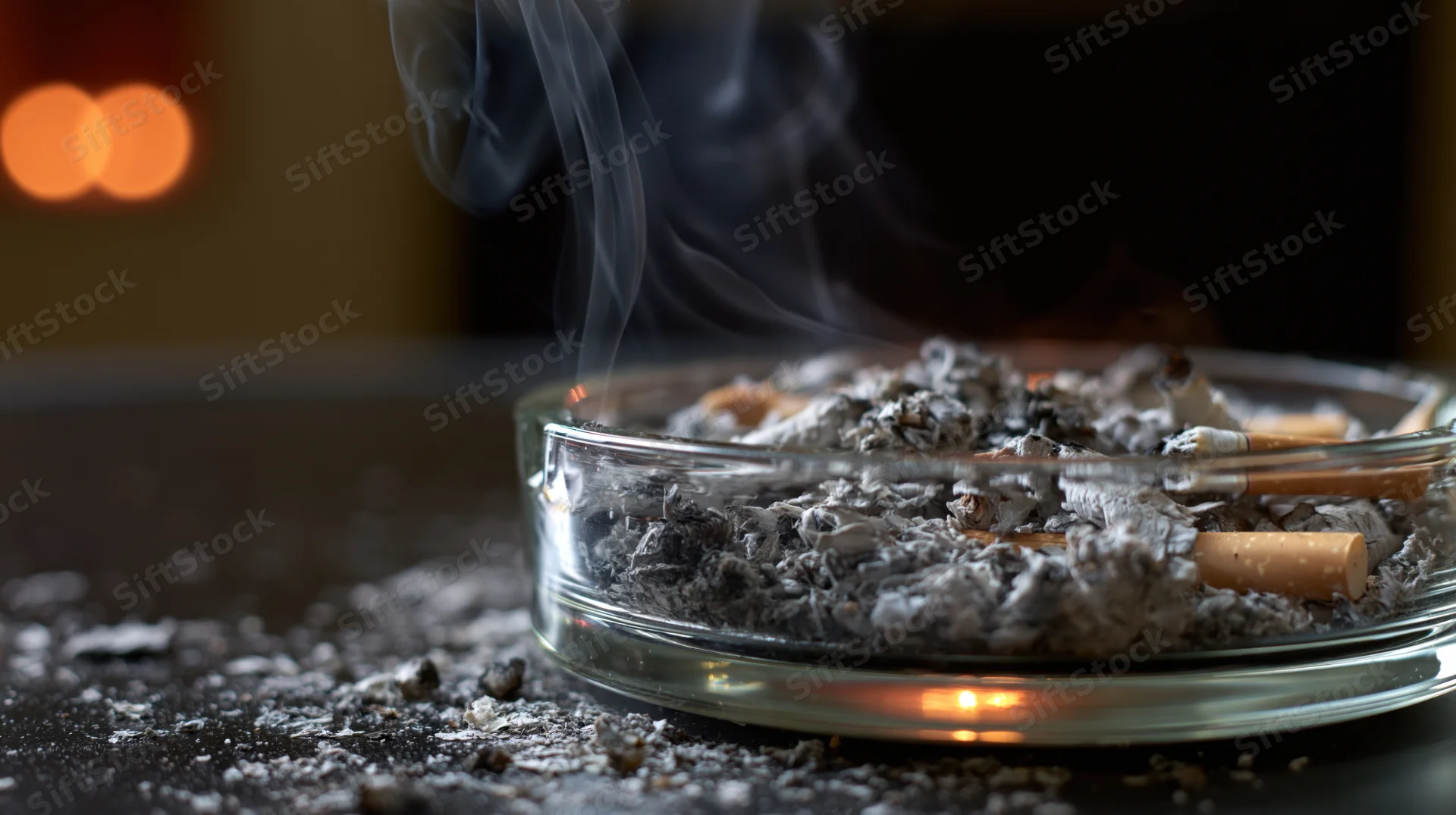 Glass ashtray overflowing with cigarette butts and ash, smoke rising, on dark surface