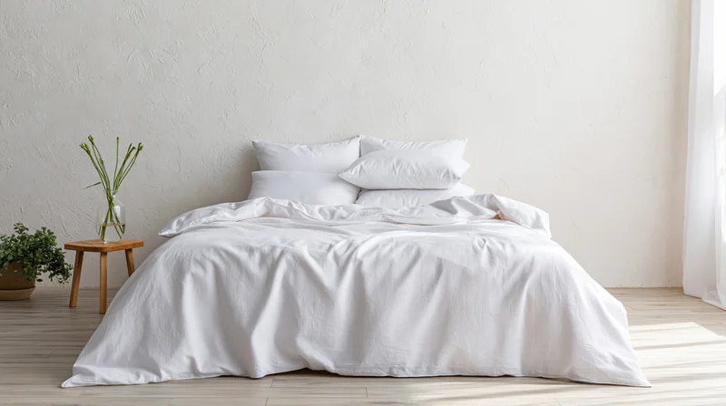 White bed with crumpled linen bedding in minimalist bedroom with wooden stool and potted plant near window