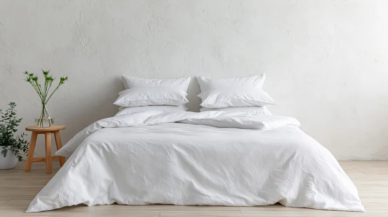 Neatly made white bed with pillows and duvet in minimalist bedroom with wooden stool and plant