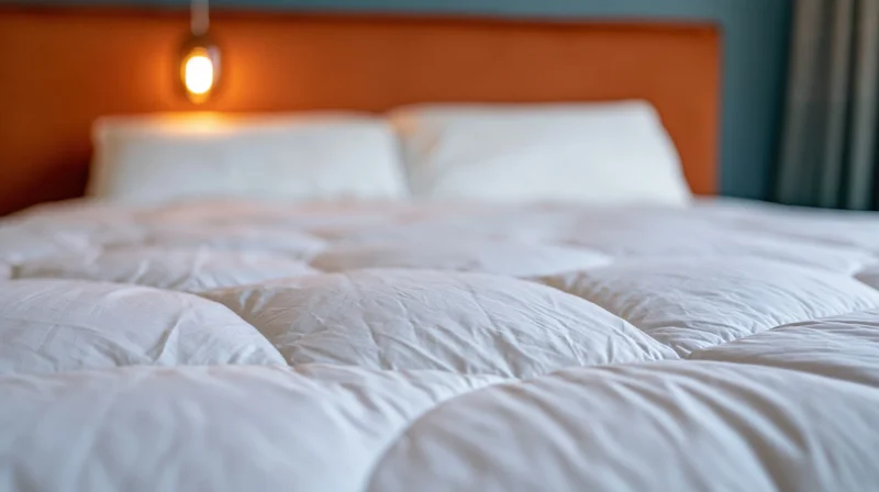 Neatly made hotel bed with white quilted duvet and two white pillows against orange headboard with warm lamp lighting