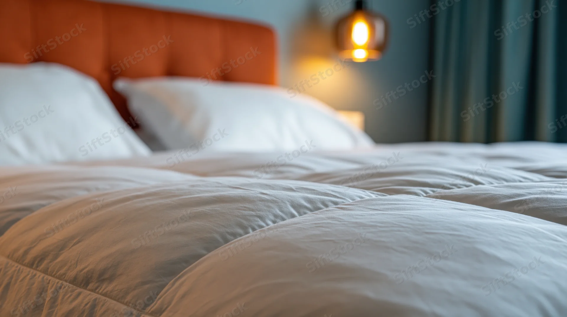 Neatly made hotel bed with white quilted duvet in foreground, orange tufted headboard, warm pendant light glowing in background