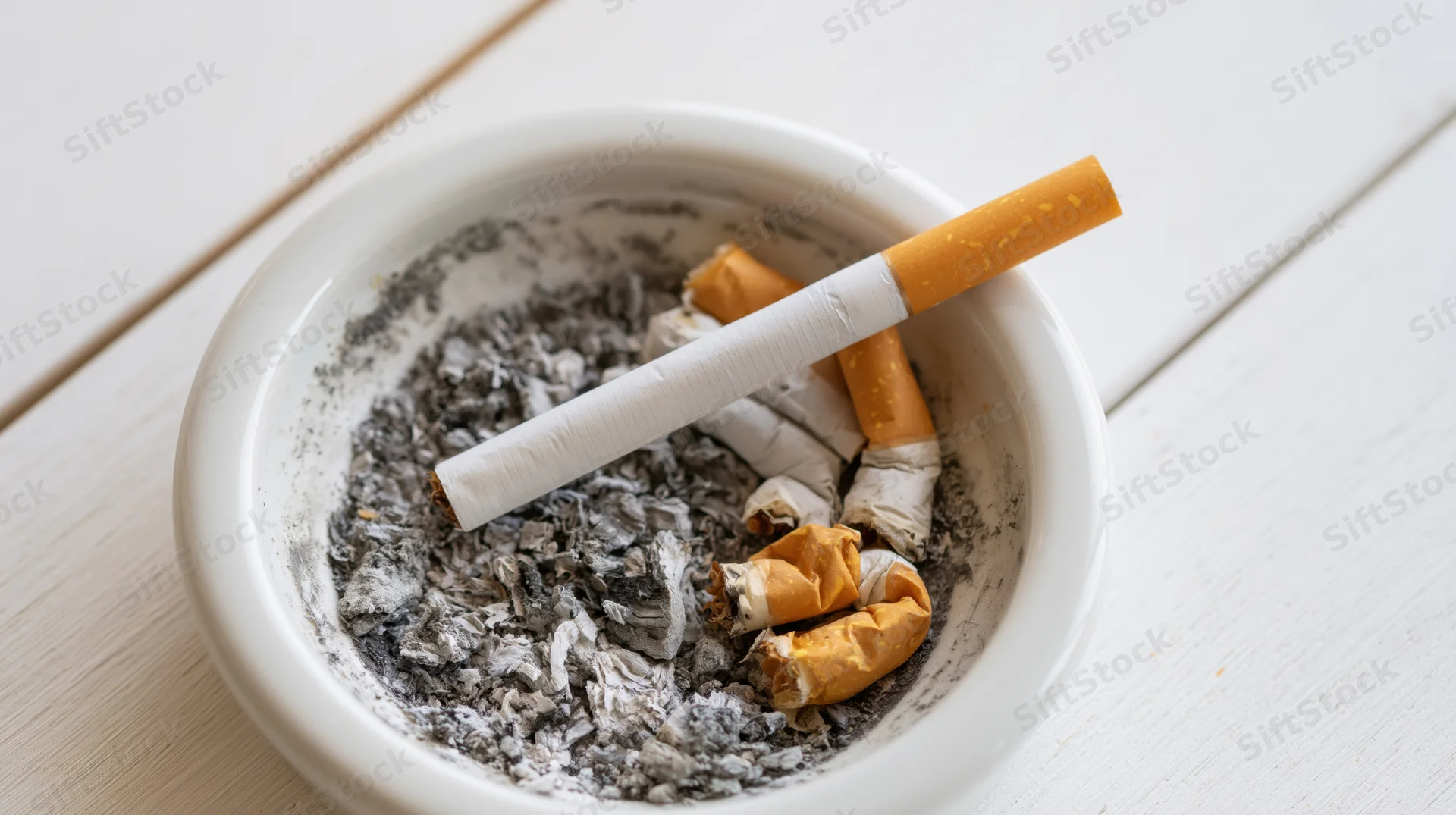 Unlit cigarette resting diagonally across a white ceramic ashtray filled with ash and cigarette butts on a wooden table