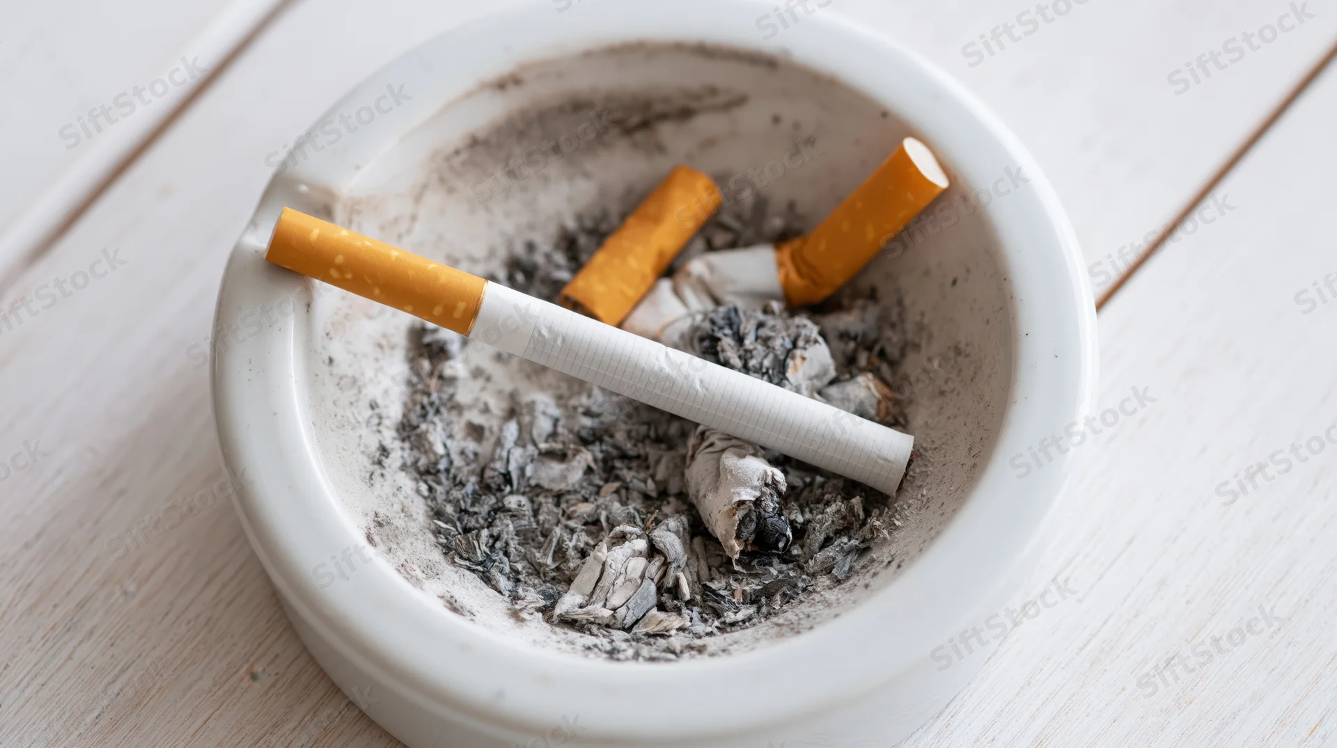 Unlit cigarette resting diagonally in a white ashtray filled with ash and cigarette butts on a wooden table