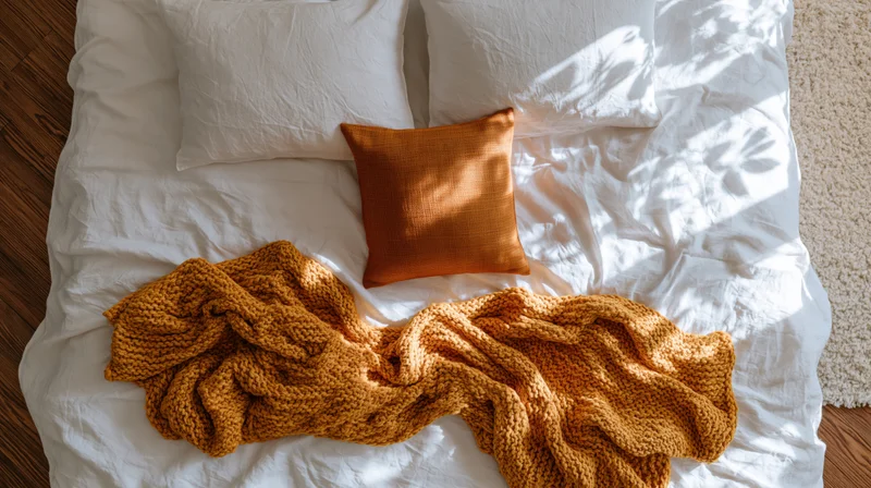 Unmade bed with white linen sheets, orange knitted blanket and pillow, overhead view in bedroom