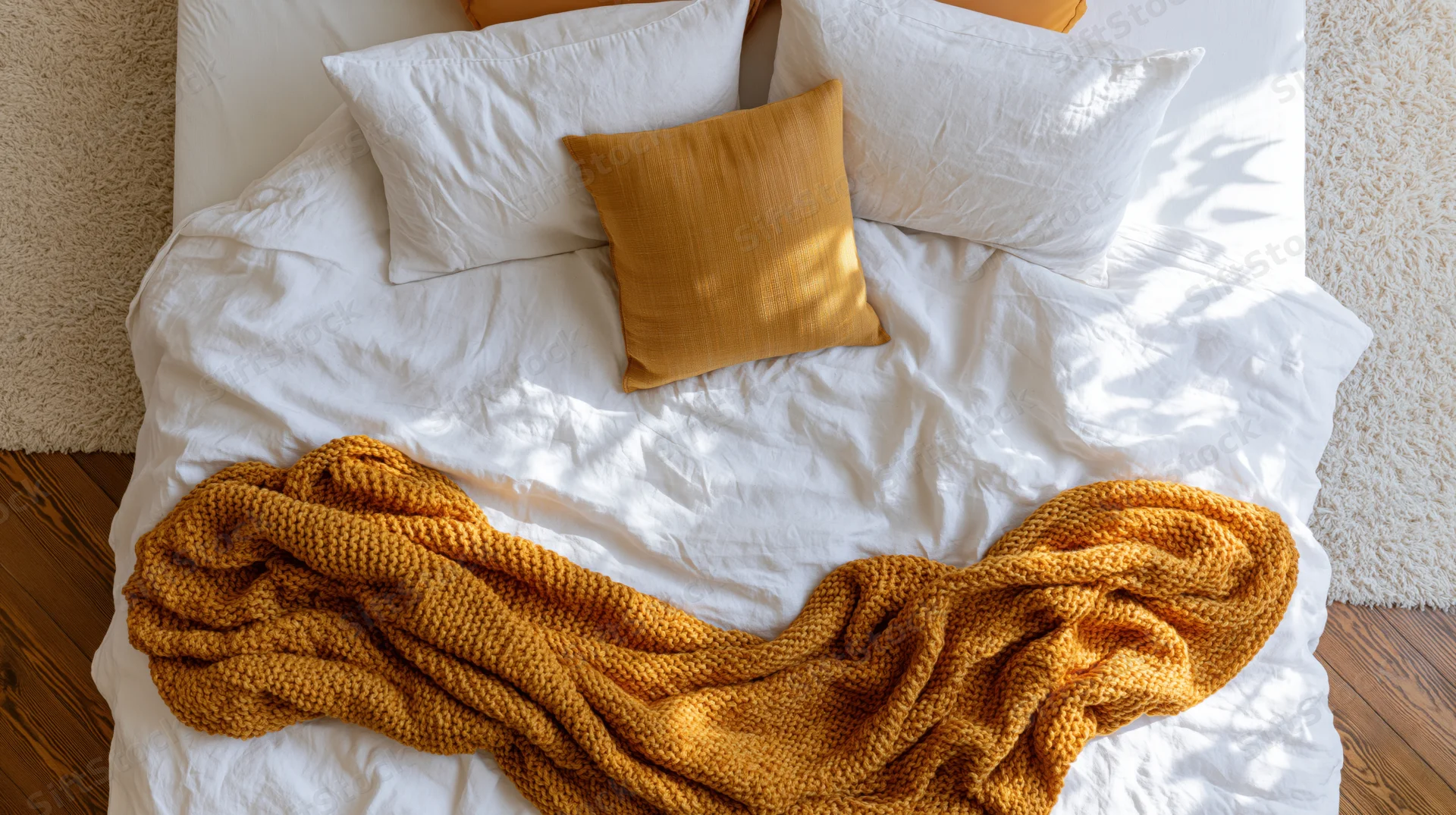 Unmade bed with white linen and mustard knitted blanket, overhead view in bedroom