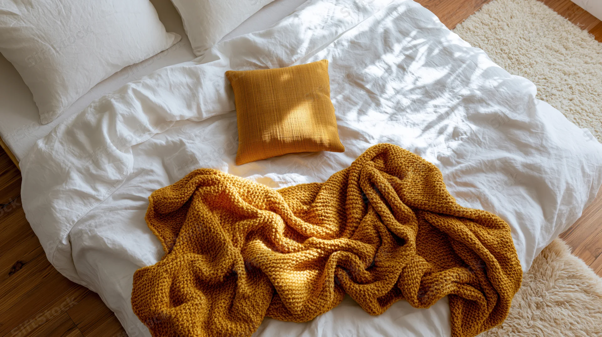 Unmade bed with white linen bedding and mustard knit blanket and pillow on wooden floor with rug