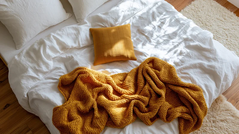 Unmade bed with white linen bedding and mustard knit blanket and pillow on wooden floor with rug