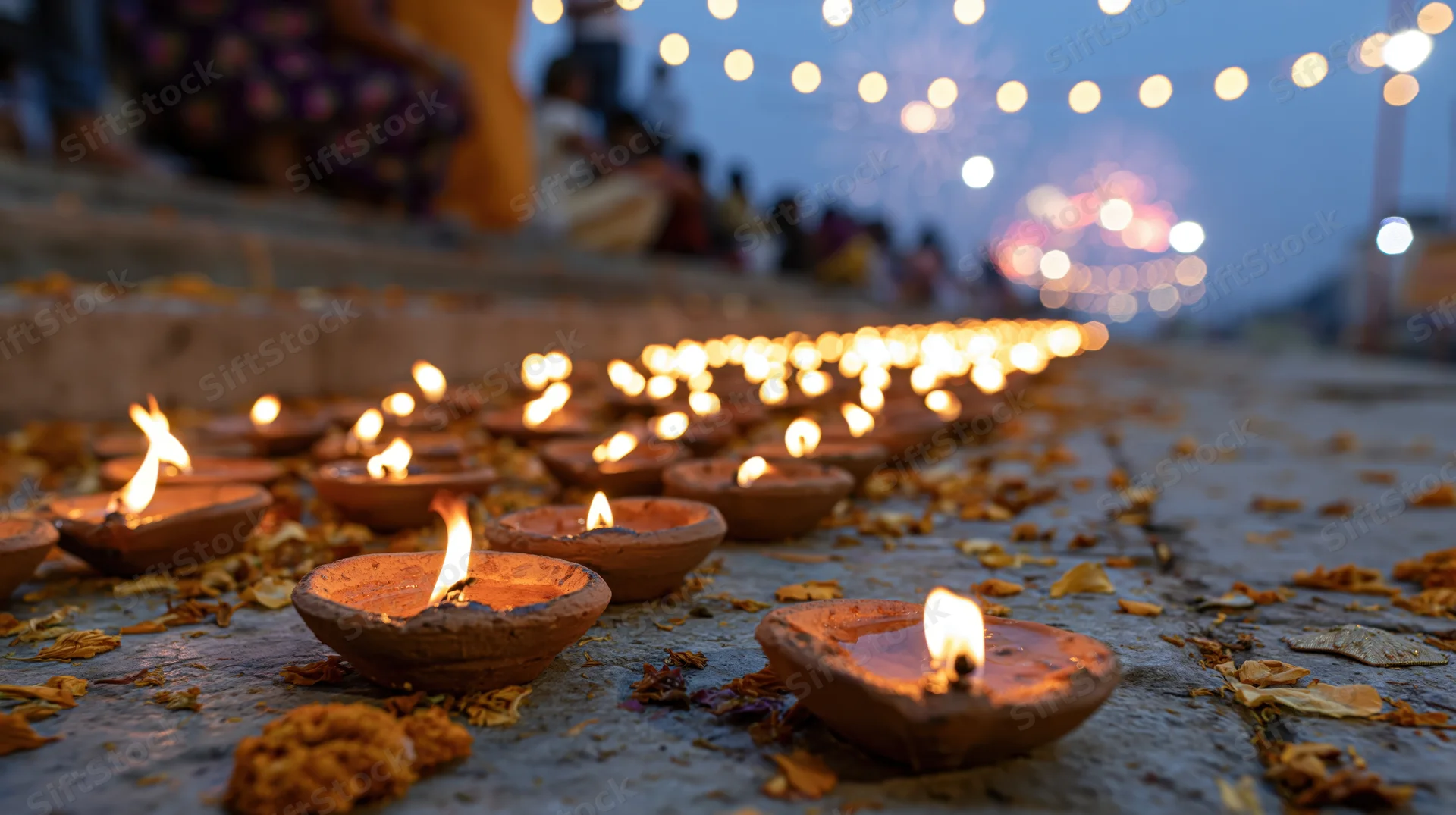 Rows of lit clay diya lamps arranged on stone ground with flower petals and string lights in background