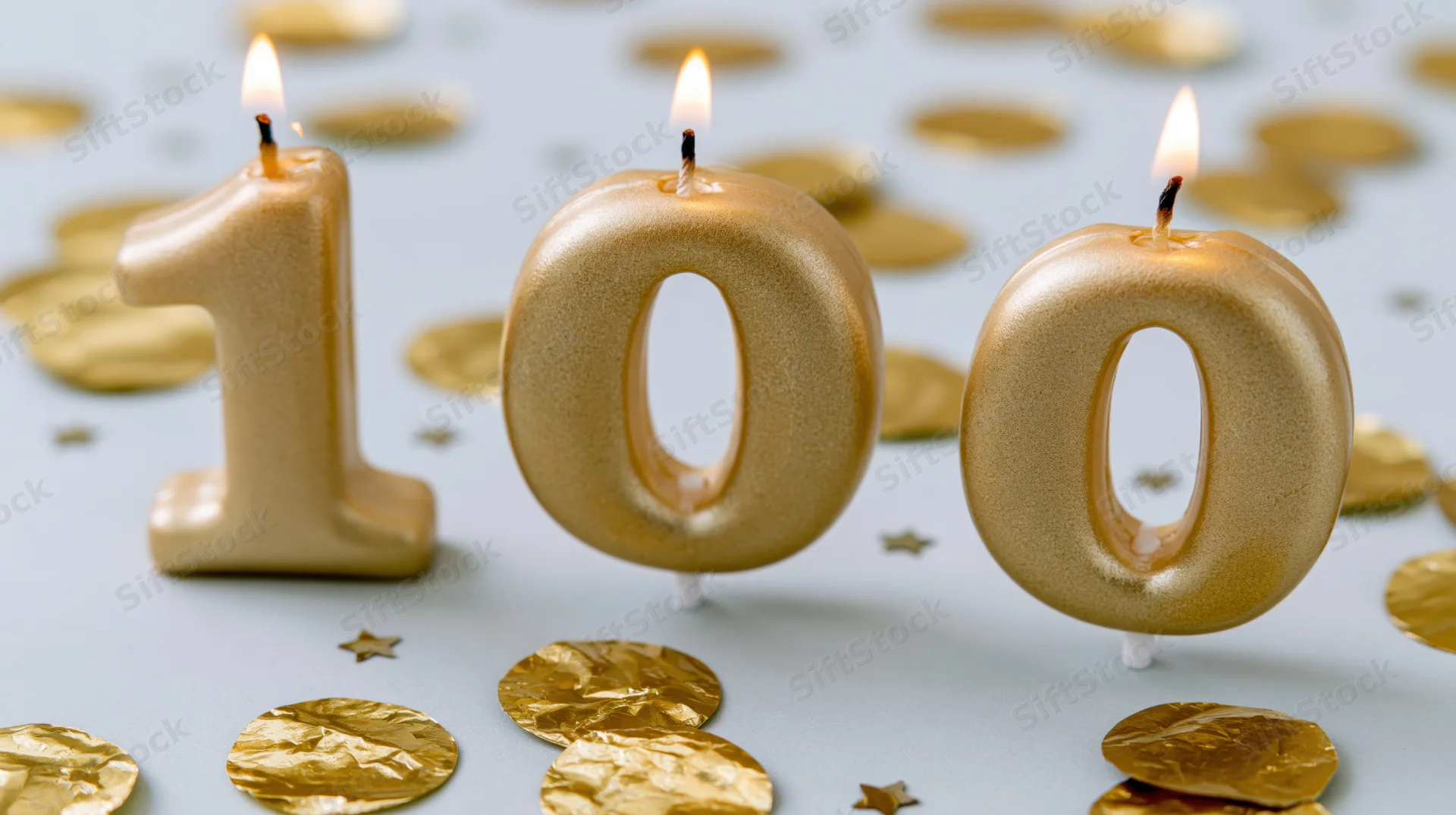 Three gold number 100 birthday candles with lit flames on a surface with golden confetti and stars