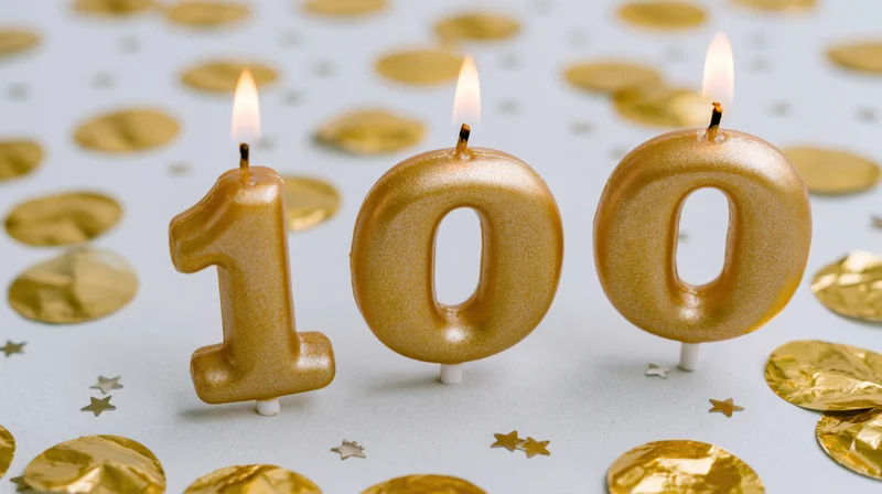 Three gold number-100 birthday candles with small lit flames