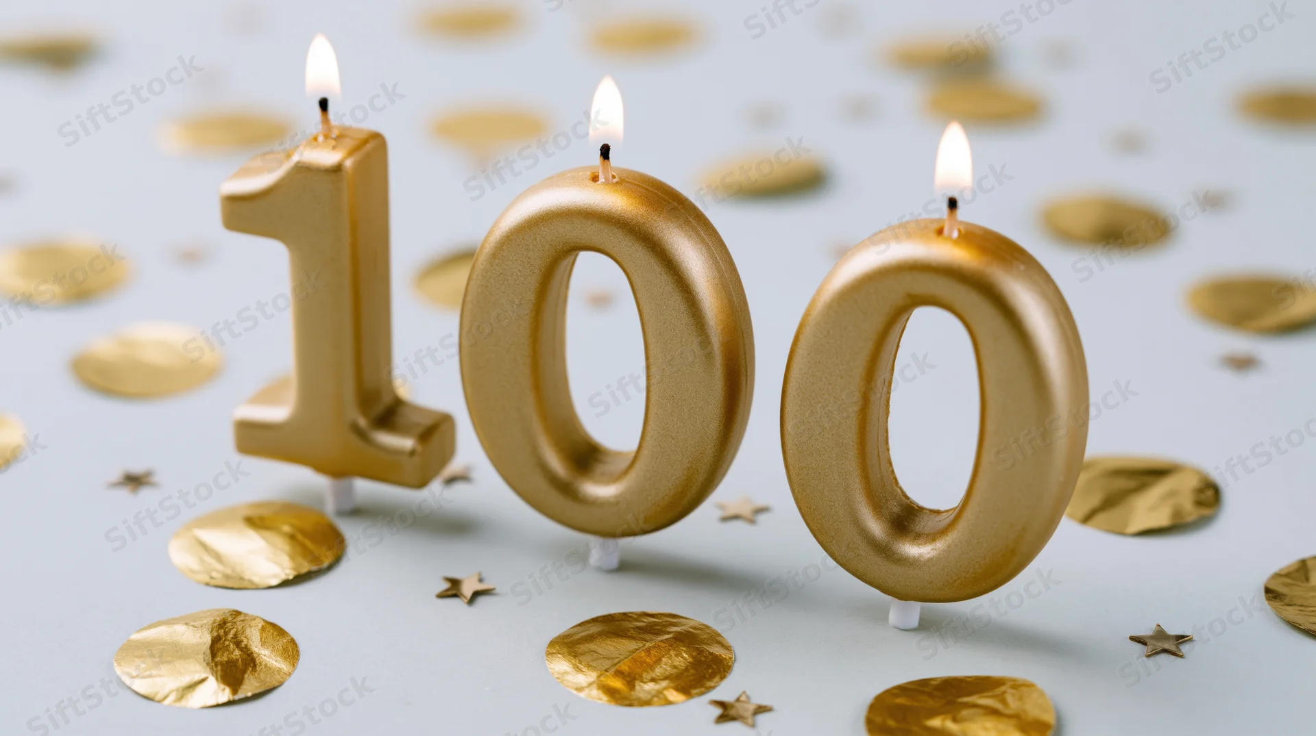 Three gold number 100 candles with lit flames on a surface with golden confetti and stars