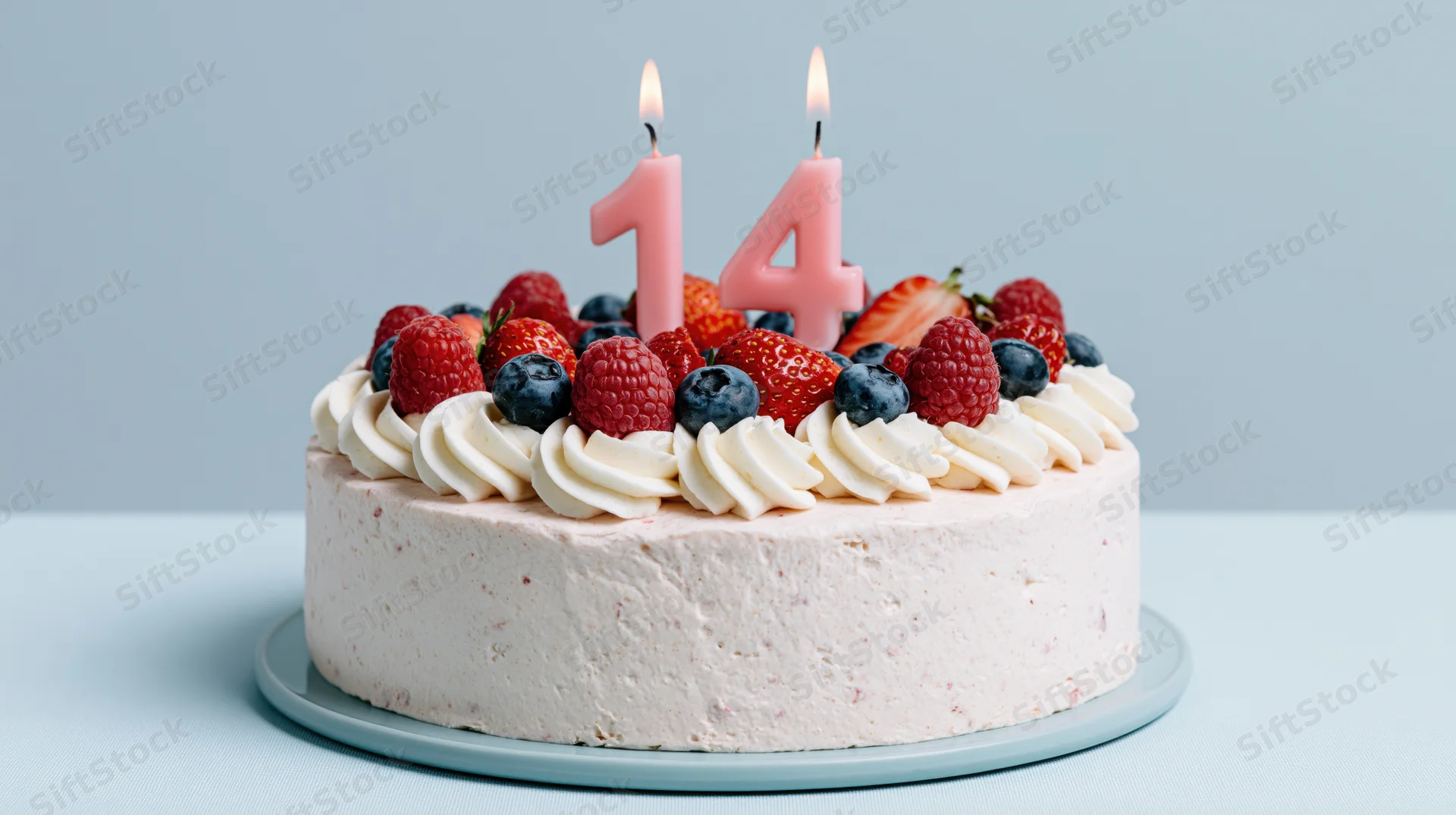 Birthday cake with lit number 14 candles and fresh berries on cream frosting, on a plate against a light blue background
