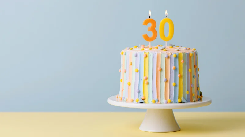 Decorated 30th birthday cake with lit number candles on white pedestal stand against blue background