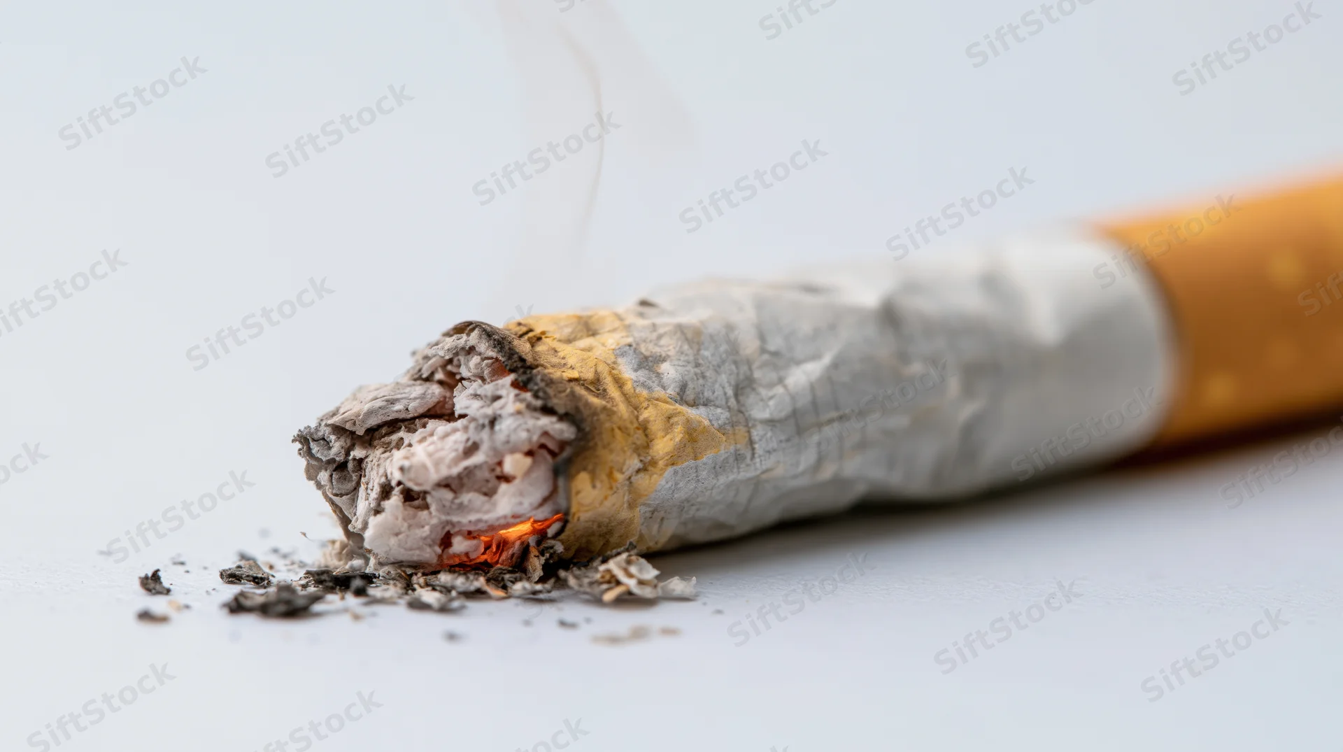 Lit cigarette butt with ash and smoke on a plain light grey surface