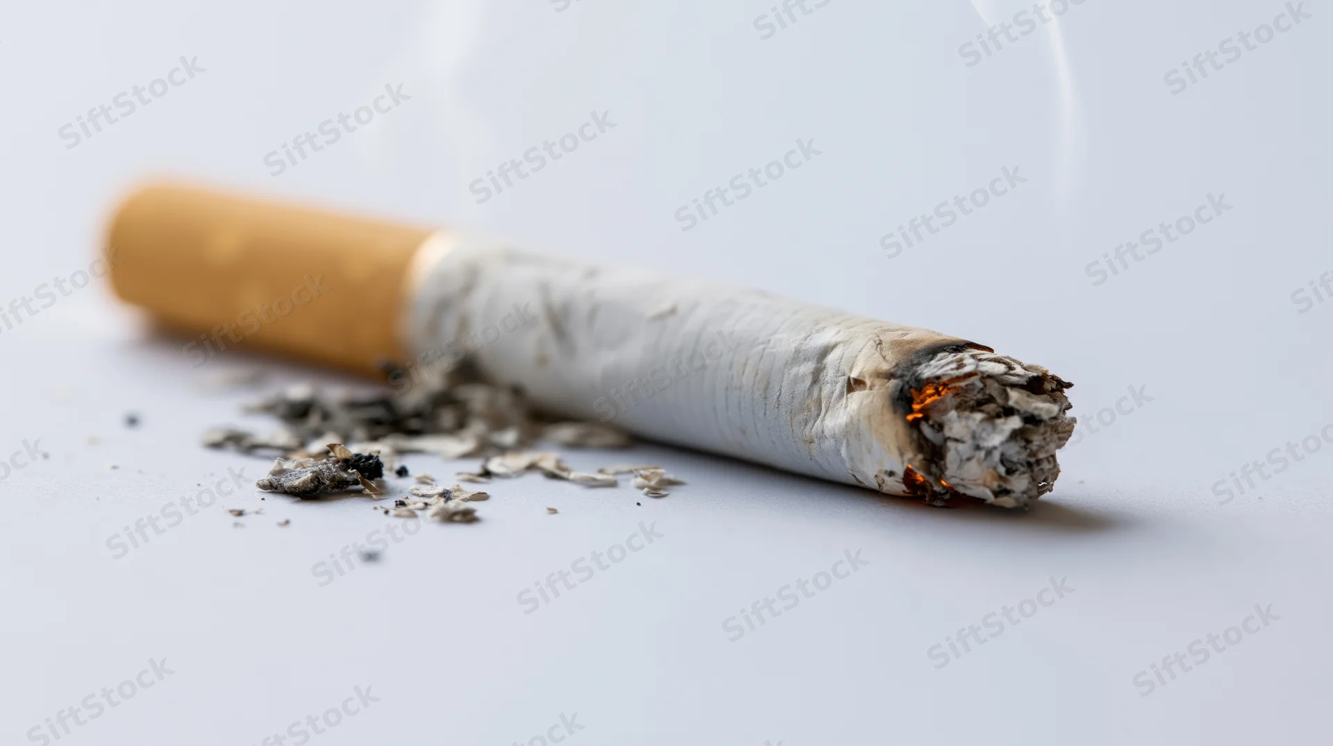 Lit cigarette butt burning on a plain light gray surface with ash and smoke