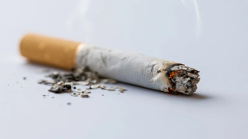 Lit cigarette butt burning on a plain light gray surface with ash and smoke