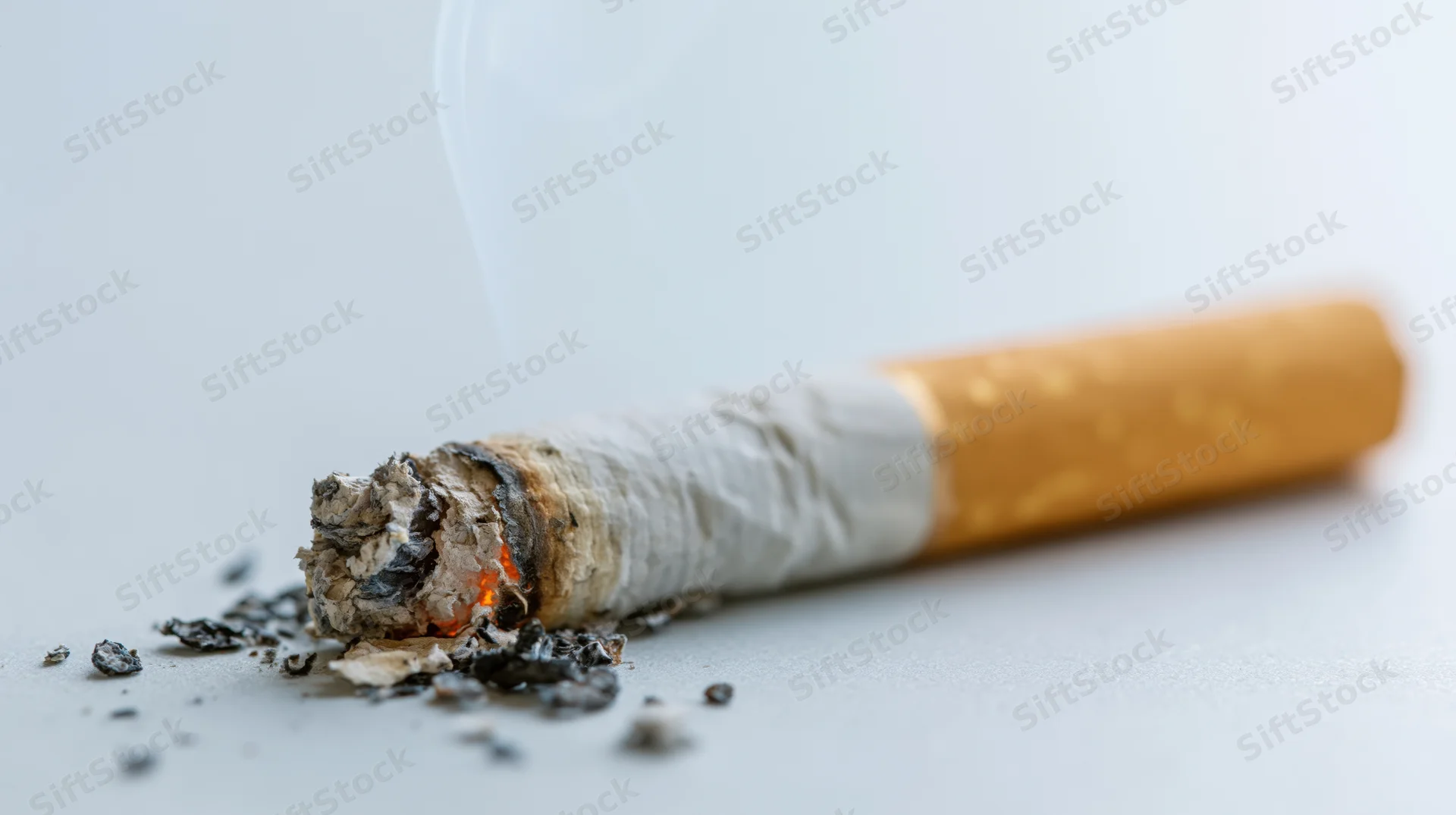 Lit cigarette butt with ash and smoke on a plain grey surface